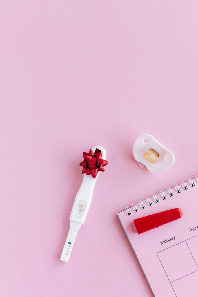 Flat lay of pregnancy test with positive result, pacifier, and calendar on pink background.