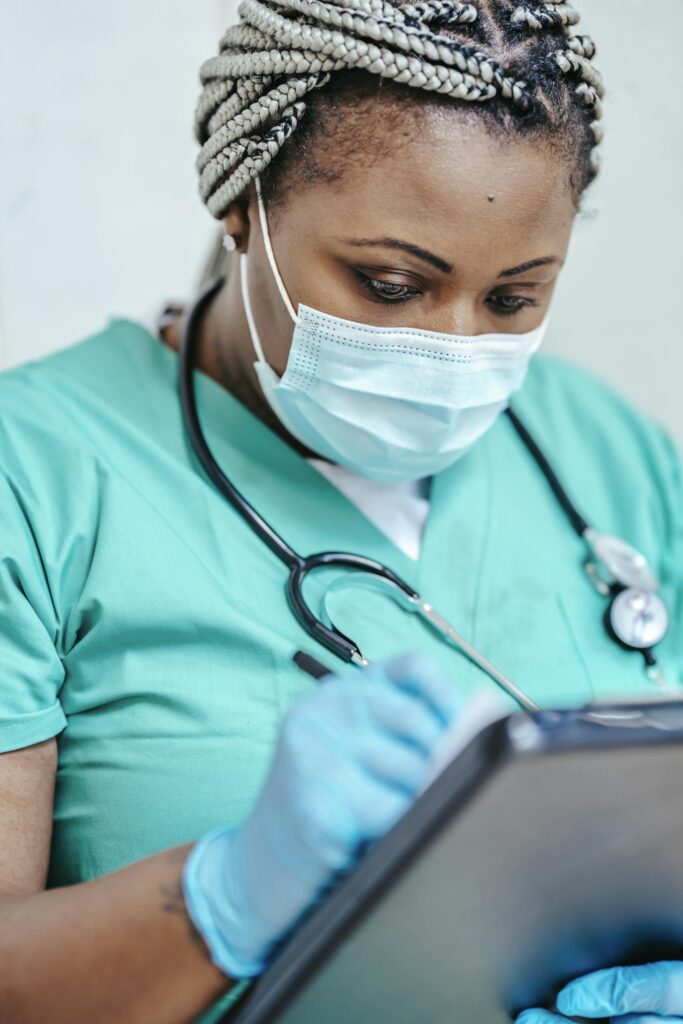 Nurse with mask and gloves reviewing documents on clipboard in a clinic setting.